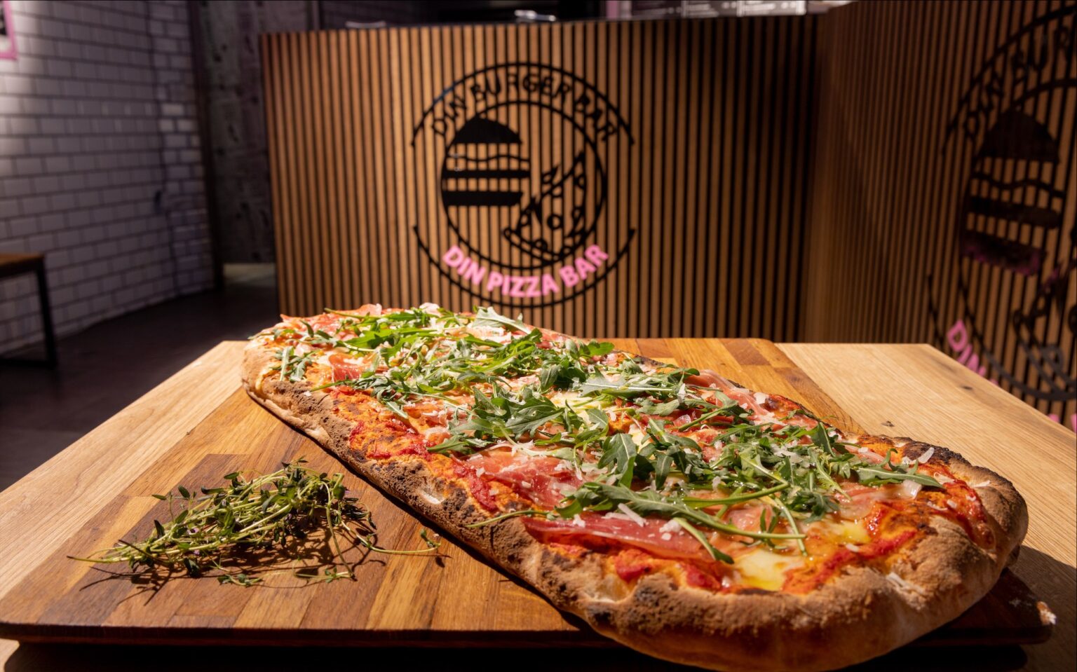 Rektangulær pizza med skinke og rucola på træbræt.